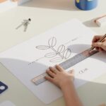 Child's hands drawing a plant cell diagram with ruler on white desk, surrounded by educational props in daylight.